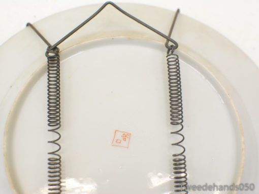 Vintage porcelain wall plate with a red mark and coiled wire hanger for decor.
